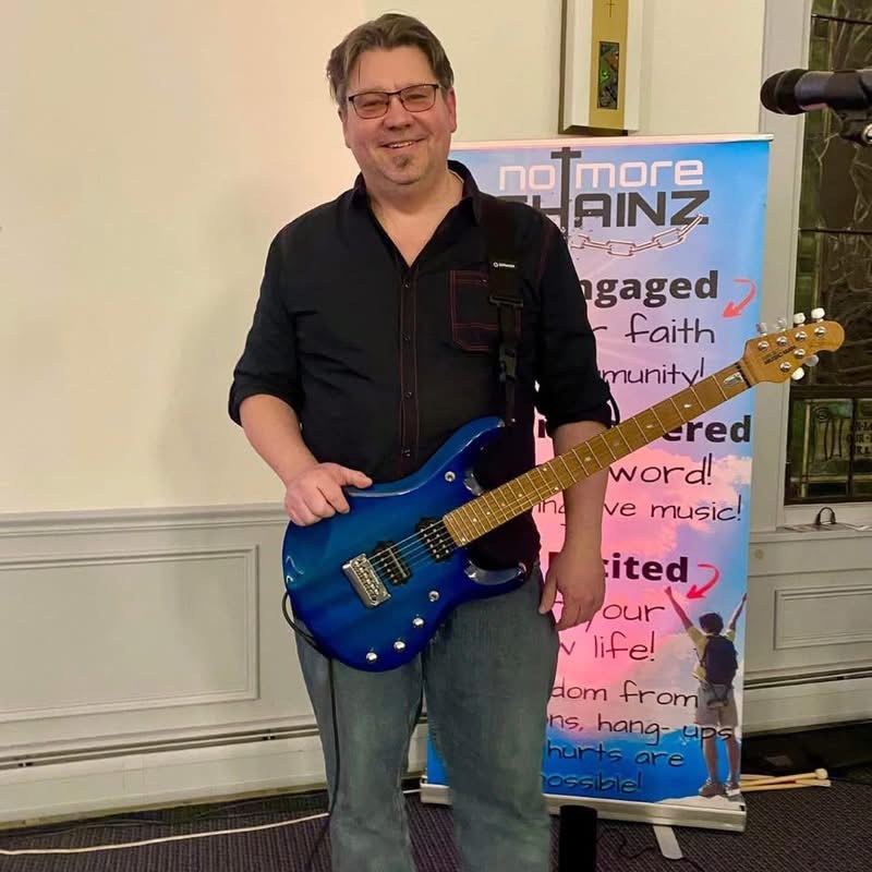 JR with blue guitar at worship beside No More CHAINZ banner